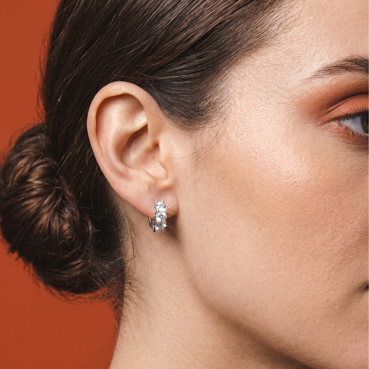 Close-up of a woman's ear wearing a silver cushion cut hoop earrings against a warm brown background