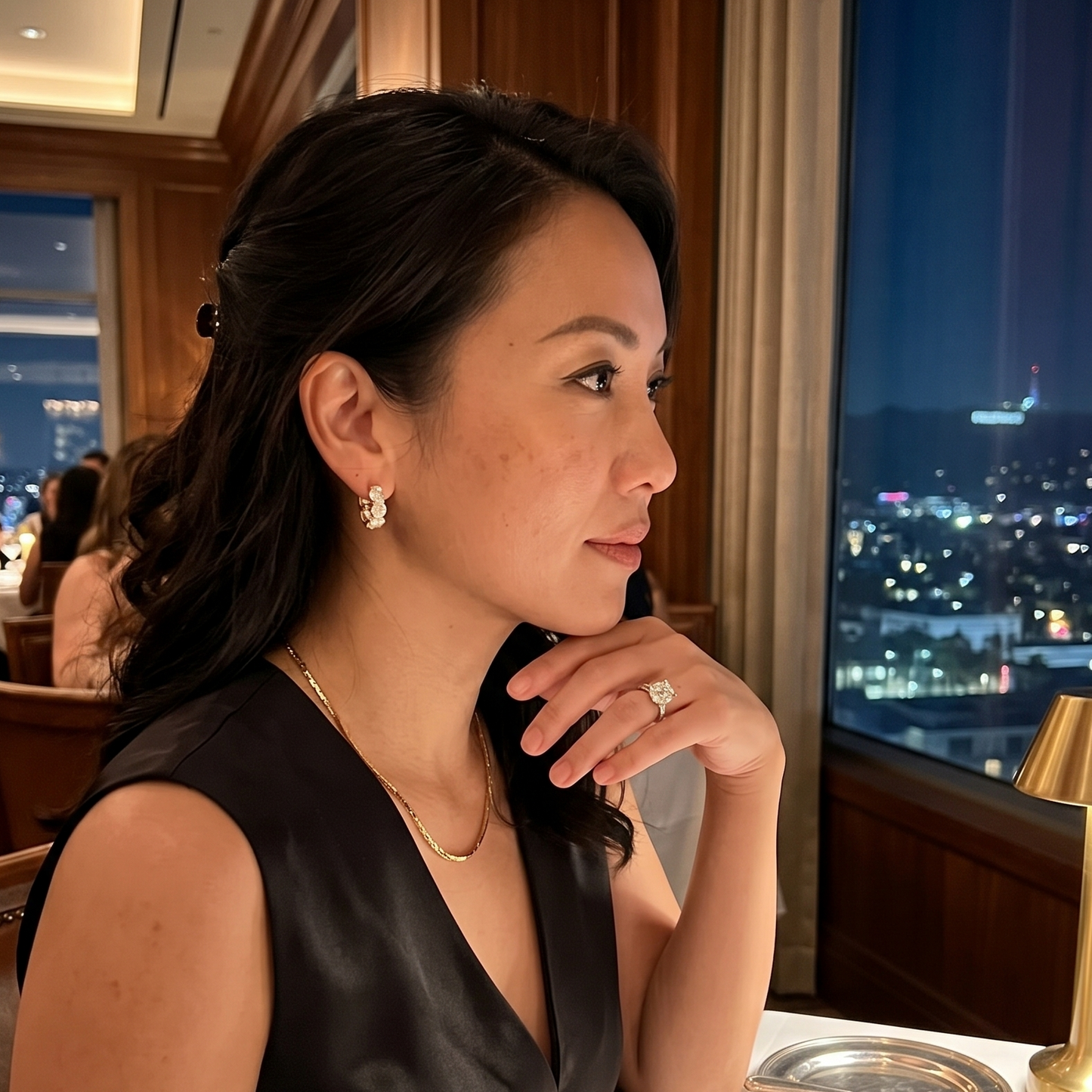 Woman wearing a gold engagement ring and oversized diamond hoop earrings sitting at a restaurant table with a cityscape view at night
